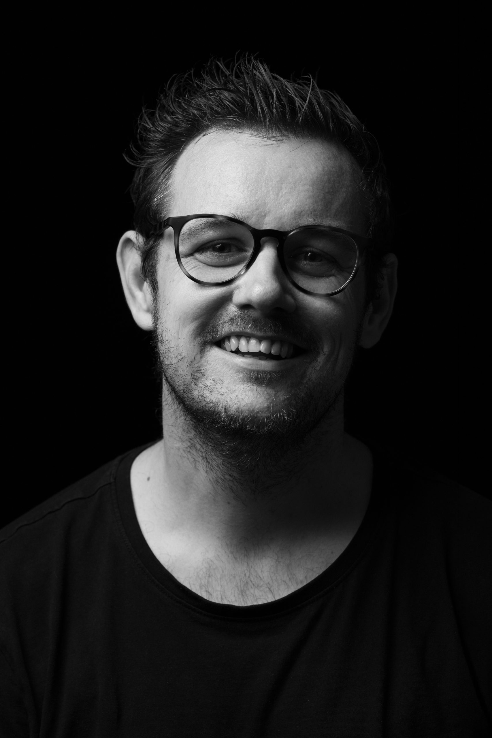 Black-and-white close-up portrait of a man with glasses smiling against a dark background, showing his eyes, nose, and teeth.